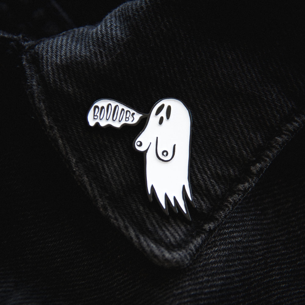 A funny horror enamel pin of a ghost with the quote, "Booobs". Pictured on a jacket lapel for unisex fashion. 