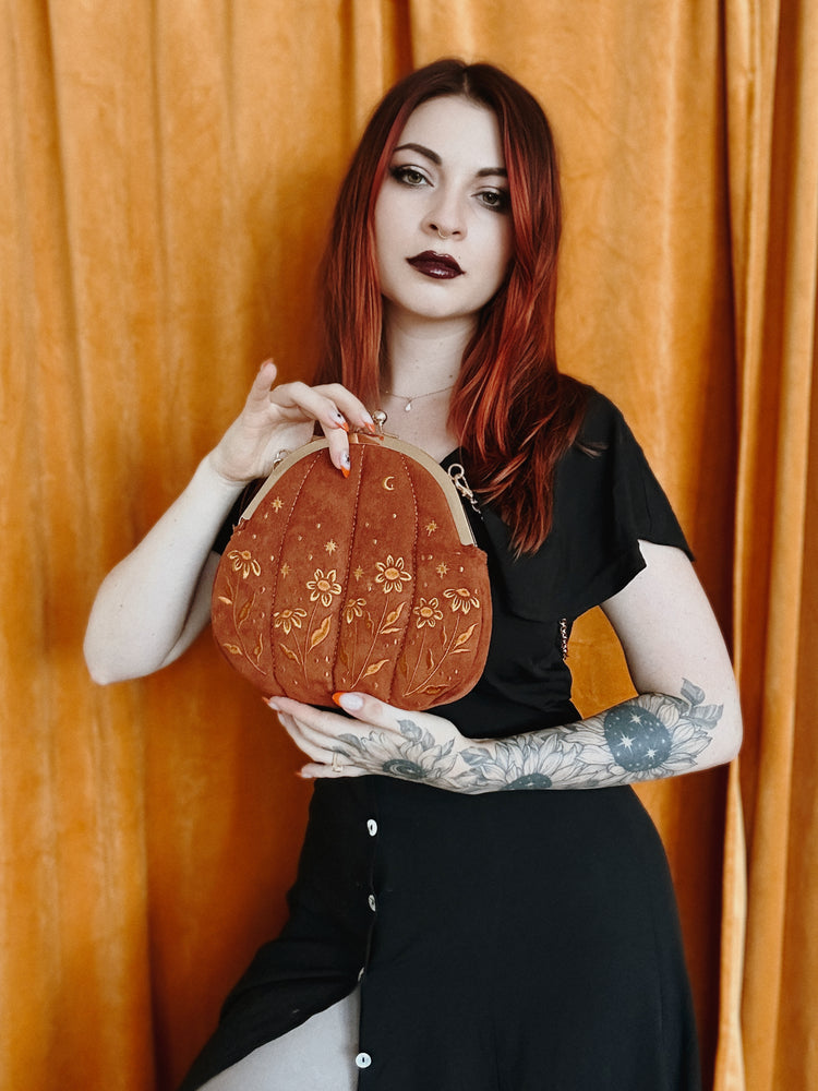 A goth style woman with black lipstick wearing a witchy pumpkin shaped purse embroidered with a botanical and celestial theme. 