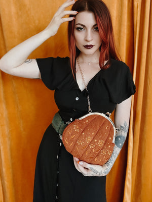 A tattooed alternative fashion woman weaing a pumpkin shaped velvet purse with a gold crossbody chain.
