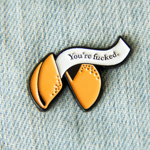 A funny enamel pin of a fortune cookie reading the quote, "You're Fucked". Pictured on a denim jacket for punk style. 