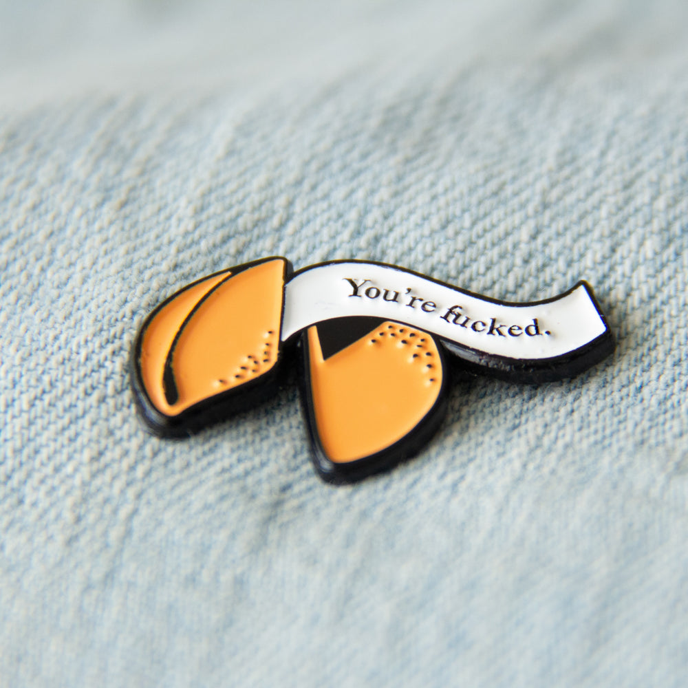 A cool enamel pin of a fortune cookie with a curse word quote. Shown on a denim vest for alternative fashion. 