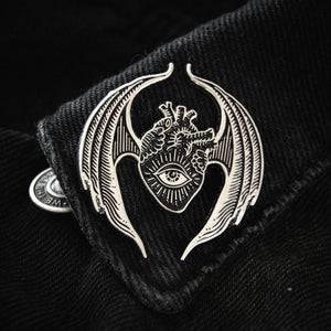 A witchy, goth aesthetic enamel pin of an anatomical heart with wings and an evil eye in black and silver. 