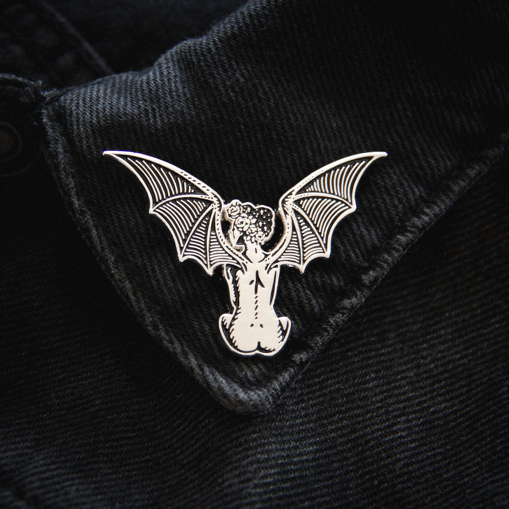 A black and silver lapel pin of a demon vampire woman with bat wings. Drawn in a style similar to Gustave Dore's woodcut art. 