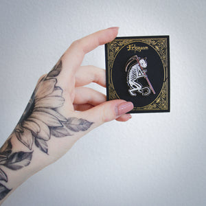 A tattooed woman holding a card with a spooky enamel pin on it of a skeleton cat holding a sickle.