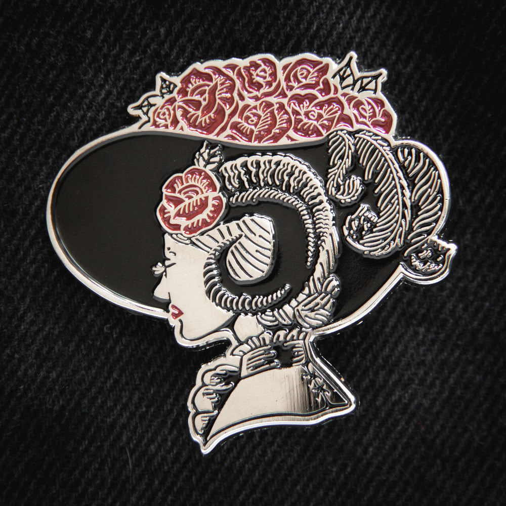 A silver, black, and red enamel pin of a Victorian woman with devil horns. She's wearing a broad derby hat with red roses. Perfect for witchy or gothic fashion. 