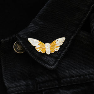 A witchy, nature themed lapel pin of a white ghost cicada, worn on a denim jacket. 