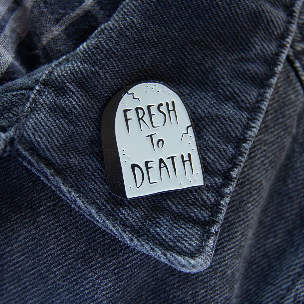 A funny enamel pin of a gravestone with the quote, "Fresh to Death", worn on a black denim jacket for alternative fashion. 