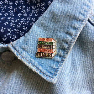 A librarian style brooch of a stack of books with the quote, "I Have Lived A 1,000 Lives". Pinned on a denim jacket lapel. 