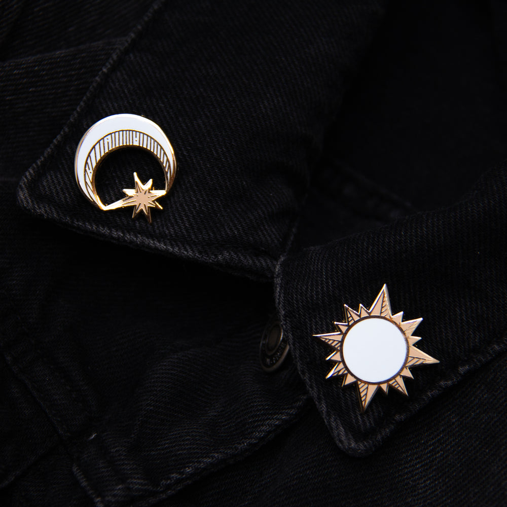 Two lapel pins worn on the collar of a denim jacket. One pin is of the moon and the other is of the sun. 