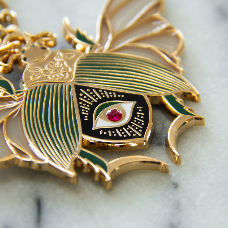 Details on a gold, black, and green purse charm of a beetle with an all seeing eye and ruby gemstone. 