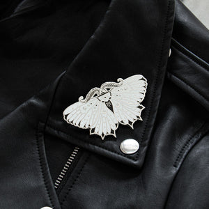 A large, witchy moth enamel pin in silver, black, and white, on the lapel of a leather jacket for cool alternative style. 