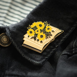 An enamel pin of a library book with yellow flowers. 