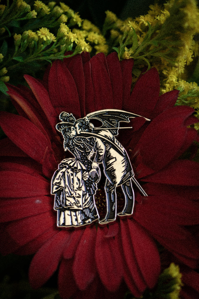 A gothic enamel pin of a victorian woman embracing a man with devil's wings. 