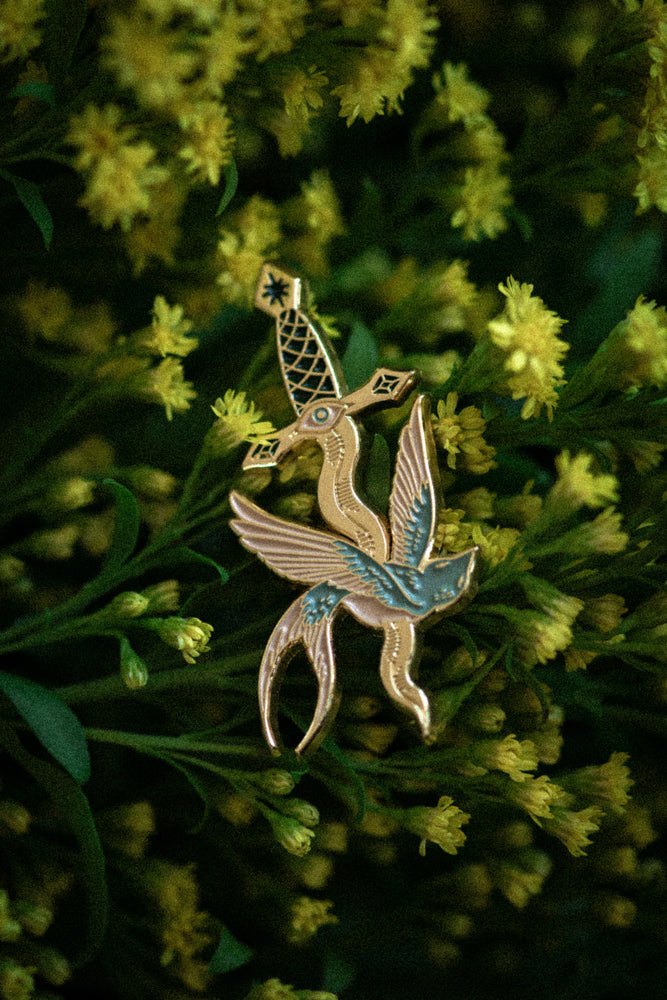 An enamel pin of a sparrow with a dagger through it, inspired by tattoo flash art. 