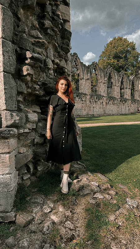 A tattooed, alternative fashion girl with red hair wearing a casual, black midi dress with vintage cape sleeves.