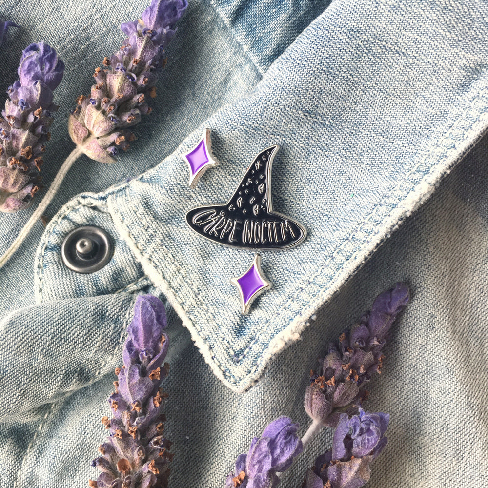 A black witch hat pin on the lapel of a denim jacket surrounded by lavender flowers. 