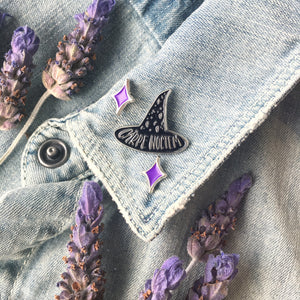 A black witch hat pin on the lapel of a denim jacket surrounded by lavender flowers. 
