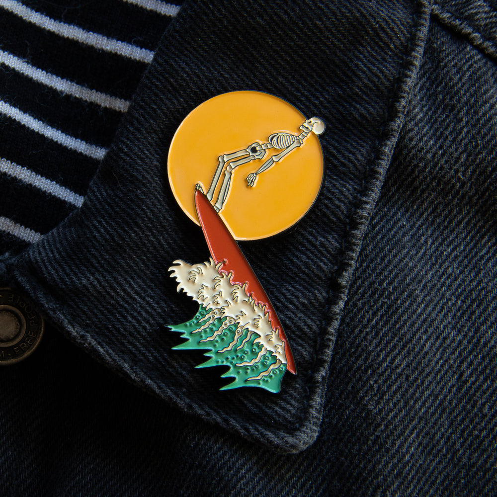 A lapel pin of a skeleton riding a surfboard in front of a beach sunset. 