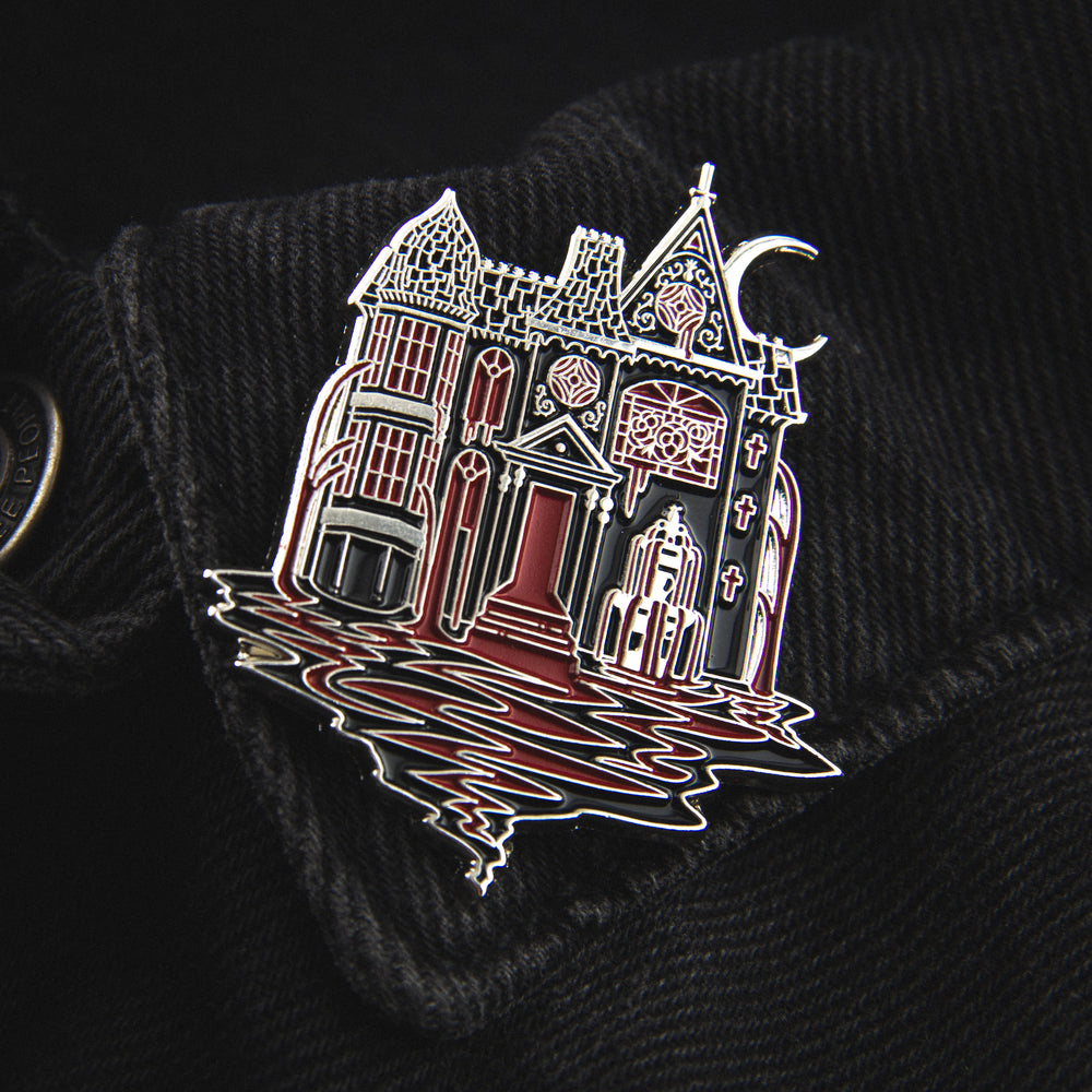 A gothic lapel pin of a haunted mansion with a silver moon behind it. 