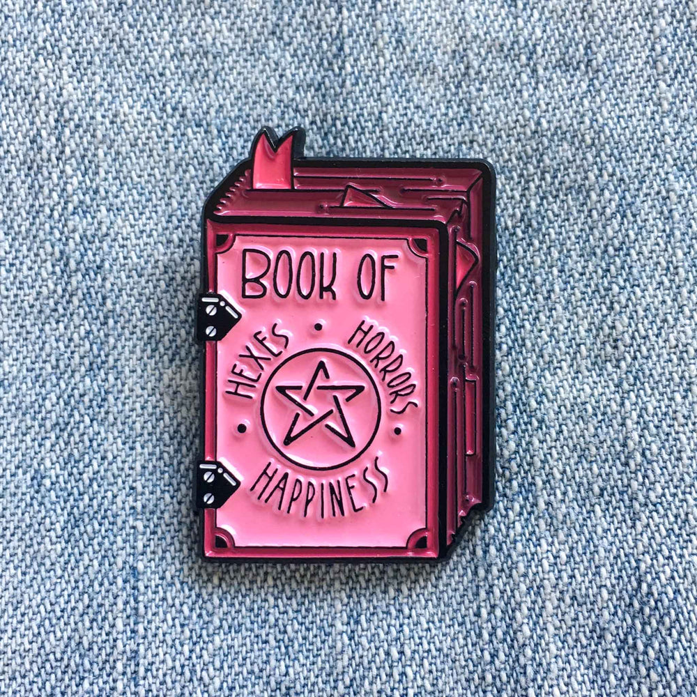 A creepy cute enamel pin of a witch's grimoire with the title, "Book of Hexes, Horrors, and Happiness". 