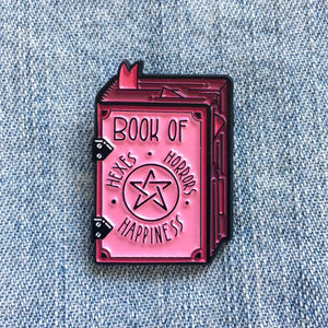 A creepy cute enamel pin of a witch's grimoire with the title, "Book of Hexes, Horrors, and Happiness". 