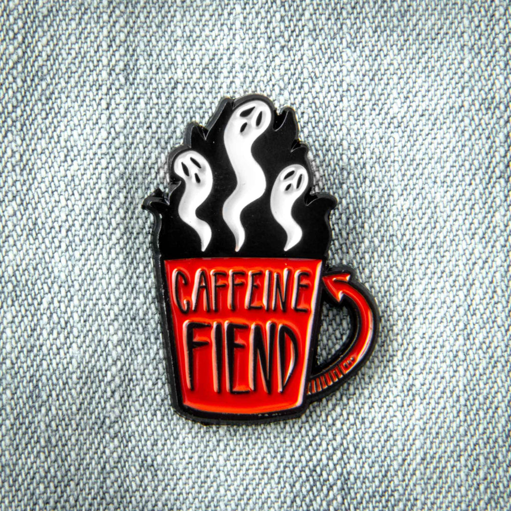 A cool enamel pin of a coffee cup with the quote, "Caffeine fiend". Ghosts rise from its brim. 