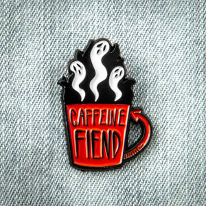 A cool enamel pin of a coffee cup with the quote, "Caffeine fiend". Ghosts rise from its brim. 