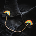 An enamel collar pin set connected with a gold chain, worn on the lapel of a denim shirt. Features two rainbows for LGBT pride. 