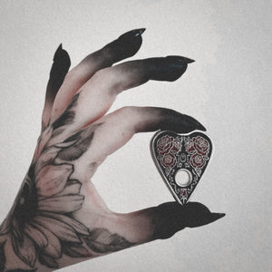 A witchy, tattooed hand holding a black, silver, and red gothic style brooch of a ouija planchette.