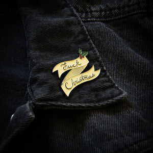 A "Fuck Christmas" enamel pin on the lapel of a denim jacket for unisex style. 