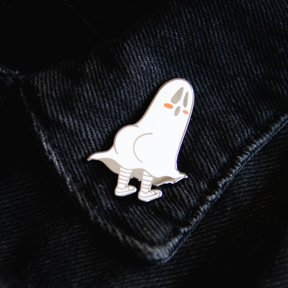 A funny black and silver spooky ghost brooch on the lapel of a black denim jacket for unisex alternative fashion.