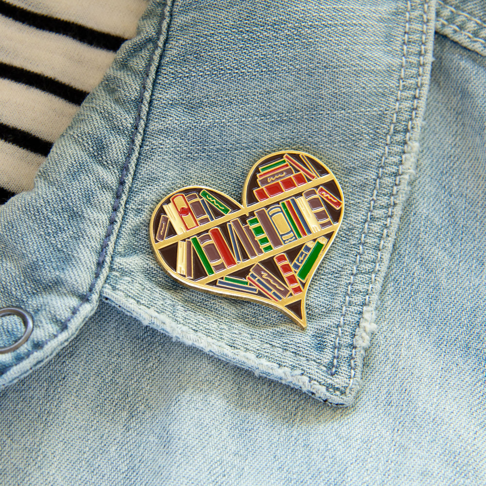 A large enamel pin of a bookshelf shaped like a heart. 