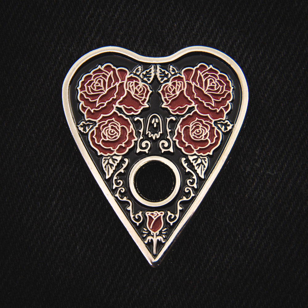 A beautiful gothic enamel pin of a ouija board planchette with red roses, filigree, and a ghost. 