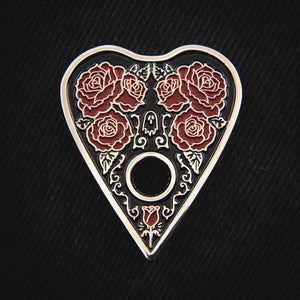 A beautiful gothic enamel pin of a ouija board planchette with red roses, filigree, and a ghost. 