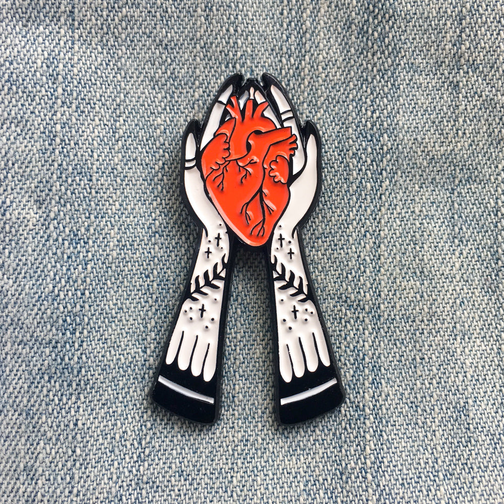 A spooky enamel pin of two ghostly hands holding a red anatomical heart. It is perfect for witchy, goth fashion. 