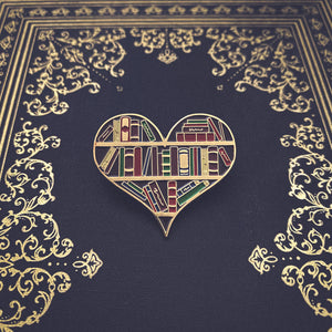 An enamel pin of a heart shaped bookshelf pictured on an antique book for dark academia fashion.