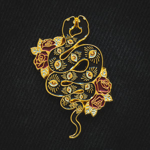 A witchy enamel pin of a gold and black snake covered in eyes for spiritual art. Red roses are on either side of the snake. 