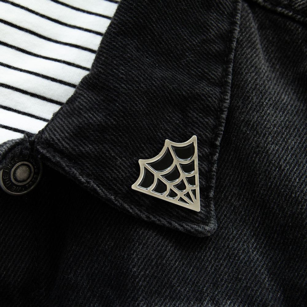 An enamel pin of a silver and black cobweb on the lapel of a denim jacket for witchy style. 