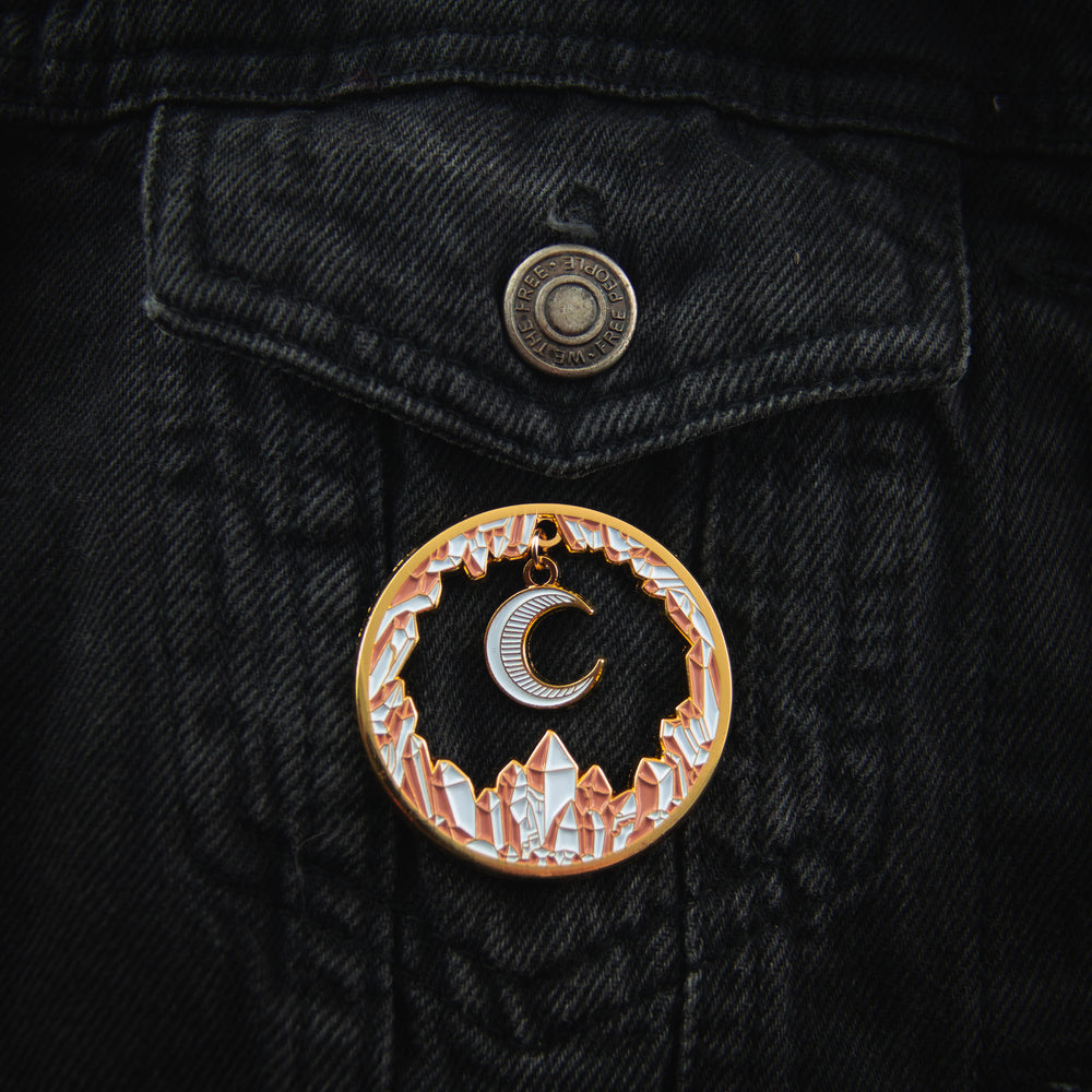 A large enamel pin of a moon charm surrounded by white and pink crystals for magical fashion. Pictured on a women's denim jacket.