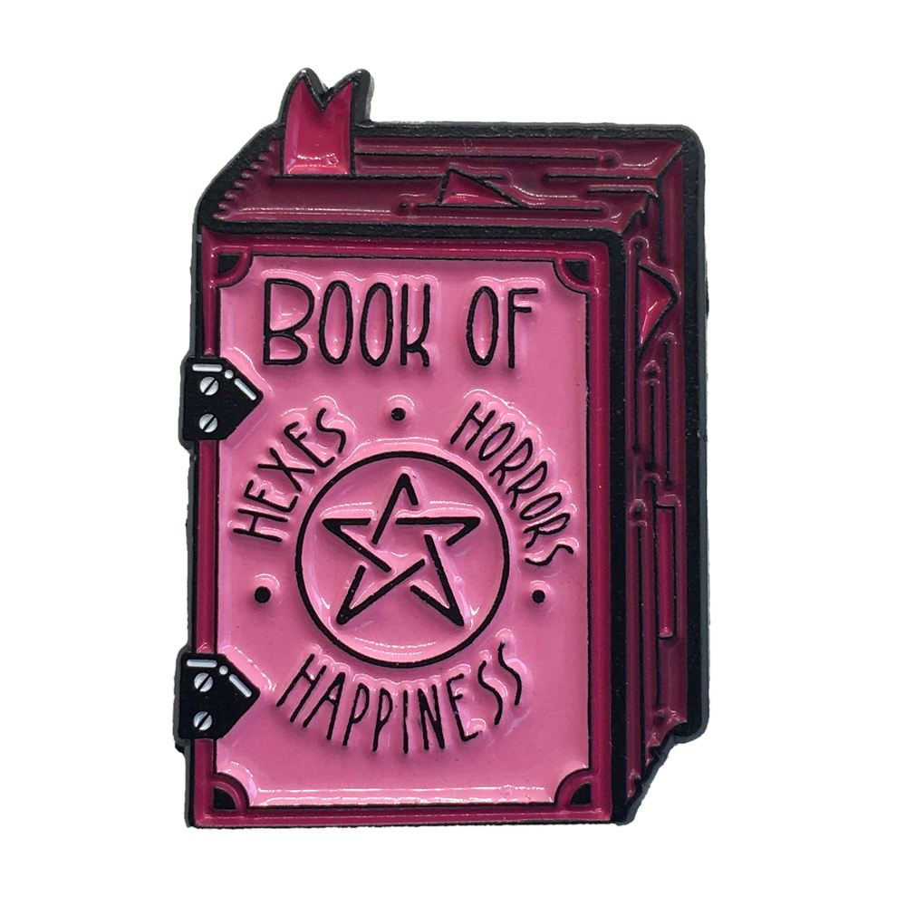 A funny black and pink pastel goth enamel pin of a spell book that says, "Hexes, Horrors, and Happiness".