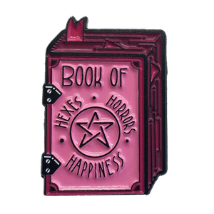 A funny black and pink pastel goth enamel pin of a spell book that says, "Hexes, Horrors, and Happiness".